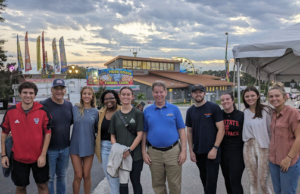 Group of students posing with director of fair in front of fairgrounds building and ferris wheel in the background.